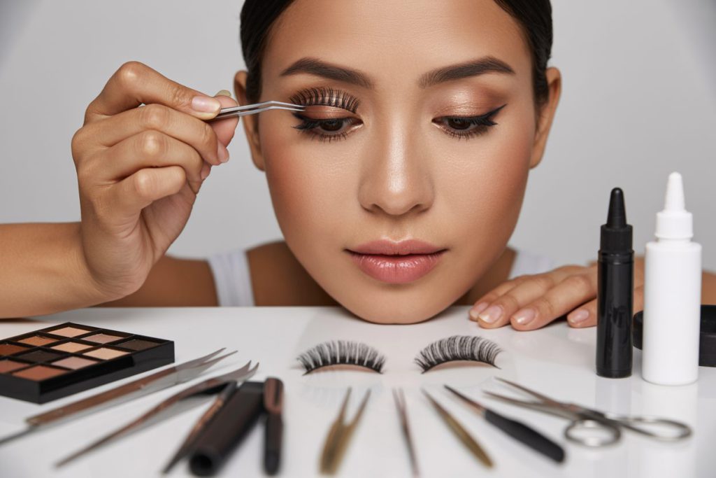 Create a realistic image of a close-up view of a young Asian female makeup artist demonstrating false lash application, with precision tweezers holding a delicate strip lash above an eye that already has perfect makeup, surrounded by professional beauty tools including lash glue, scissors, and various types of false lashes arranged neatly on a clean white vanity, with soft, flattering lighting highlighting the precision of the technique.