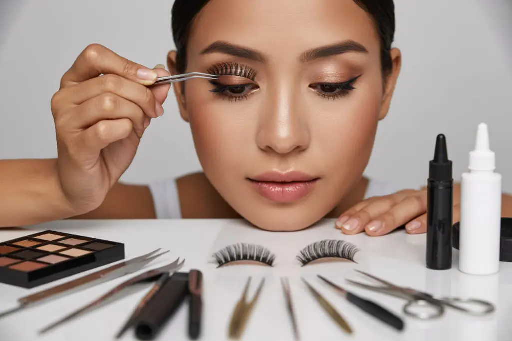 Create a realistic image of a close-up view of a young Asian female makeup artist demonstrating false lash application, with precision tweezers holding a delicate strip lash above an eye that already has perfect makeup, surrounded by professional beauty tools including lash glue, scissors, and various types of false lashes arranged neatly on a clean white vanity, with soft, flattering lighting highlighting the precision of the technique.